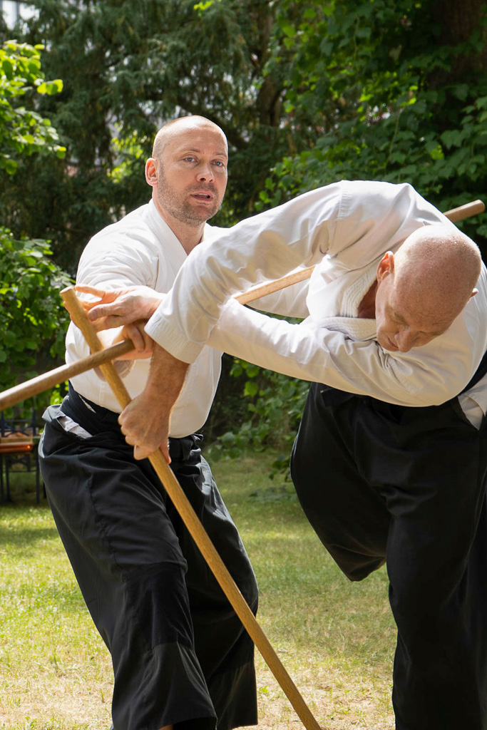 Zwei Aikidoka in traditioneller Kleidung, die eine Wurftechnik mit dem Jo praktizieren.